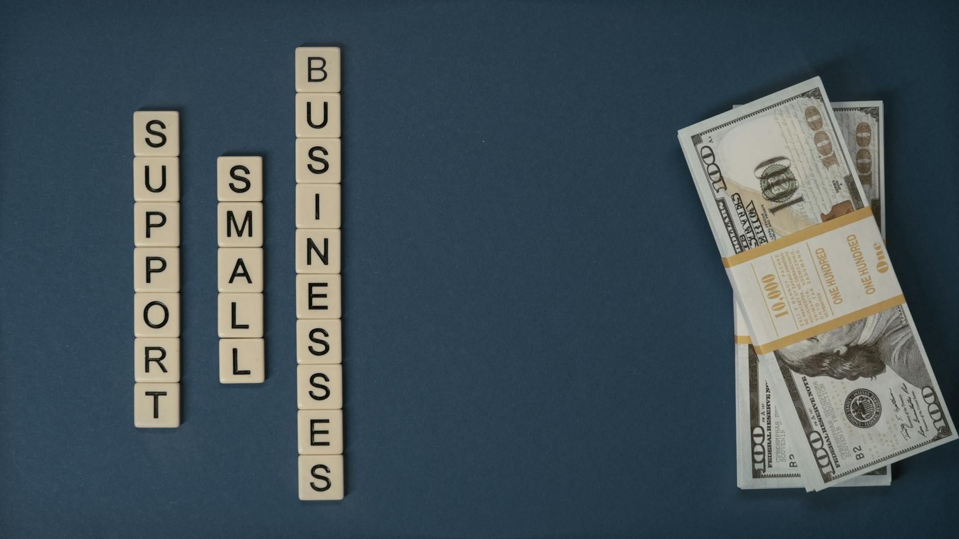 Træbrikker med teksten Support Small Businesses ved siden af en stak dollarsedler.